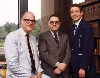 Three men sitting on a desk, posing for a photo. The left and center (Dr. Joy) men have dark rimmed glasses on. The man on the right is Dr. Dale Smith.