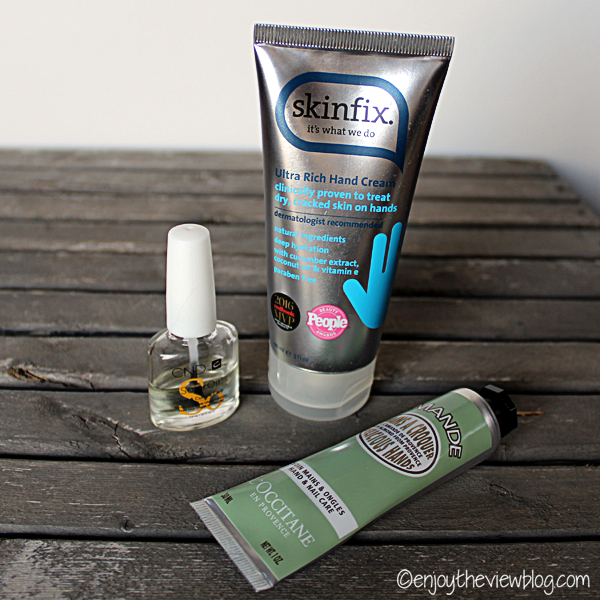 tube of Skinfix hand cream, bottle of CND Solar OIl, and tube of L'Occitane Almond hand cream lying on a wooden surface