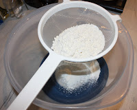 A sieve full of flour, baking powder and salt, over a large plastic bowl