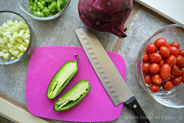 Fresh ingredients are being prepared for this flavorful corn salad. A set of small clear glass bowls filled with chopped fresh green peppers, chopped celery, whole cherry tomatoes surround a small purple plastic cutting board. A fresh jalapeno pepper is sliced in half on top of the cutting board and whole fresh purple onion sits beside the cutting board. 