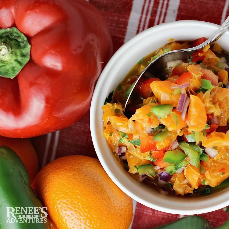 Making citrus salsa. overhead shot of salsa in white bowl with spoon, red bell pepper, jalapeno, and clementine orange off to side