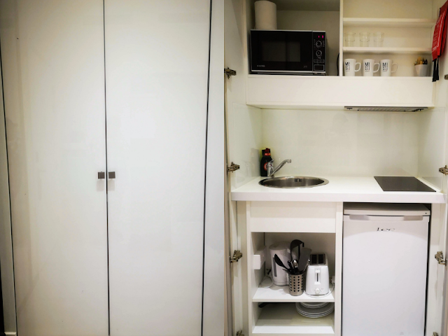 Hidden kitchenette available at Nox Kensington. The first image shows the white doors closed. The next image shows the doors open. You can see the under counter fridge, cutlery and crockery. There is a stainless steel sink and an electric 2 ring black glass hob. Above this is a microwave and a collection of cups and glasses.