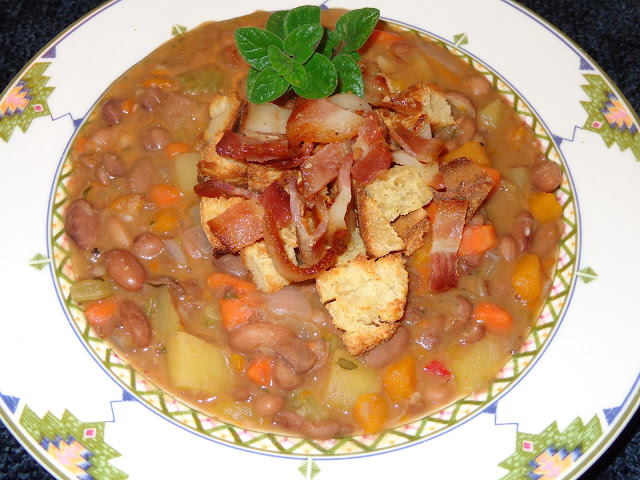 PORTIONS: 4 INGREDIENTS 12 oz. / 340 g dried pinto beans 6 cups chicken broth or water with 1 chicken bouillon without MSG 1 tsp. salt 5 slices thick bacon 1 cup red onions, diced 2 garlic cloves, minced 1 celery rib, diced ½ yellow pepper, diced ½ orange pepper, diced ½ cup red pepper, diced 1 large carrot, diced 1 cup butternut squash, diced 1 potato, diced with or without skin 2 tsp. fresh thyme leaves 3 tsp. fresh oregano leaves ¼ tsp ground pepper ½ cup croutons METHOD Check the beans for stones o any foreign objects in it. Place the beans in a pot covered with plenty water and let it soak for about 8 hours or overnight. Discard the water and fill with 6 cups of fresh water and salt. Bring it to a boil and with a ladle remove the foam from the top. Reduce the heat to simmer, cover and cook the beans for about 1 hour or until beans are very soft. Cook the bacon in a frying pan at low heat. Cook it the way you like it and set aside. Using same frying pan with the bacon fat, add garlic and onions and cook until transparent. Add and slightly brown the vegetables, scraping the bottom of the pan to lose the bacon particles. Incorporate the vegetables to the beans. Using some of the water of the beans deglaze the frying pan and add it to the beans. Add potato, herbs, water, chicken bouillon and pepper. Bring it to a boil, reduce heat, cover and let it simmer for about 15 minutes until potato is cook. Cut the bacon in small pieces. Serve the soup with croutons and bacon.  