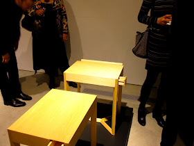 People at an exhibition opening, looking at two small wooden side tables.