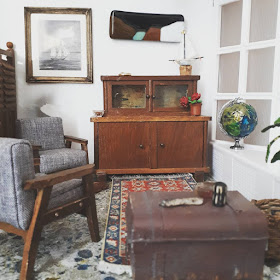 1/12 scale modern miniature lounge scene with two mid-century modern armchairs in grey with dark stained arms and legs, an early twentieth century cupboard, a battered sea chest and afghan rug on the floor. On the wall is a framed picutre of a boat and an abstract landscape. On the windowsill is a globe. 