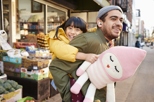 A man carrying a small girl on his back on a busy sidewalk, he has Pixel Buds in his ears