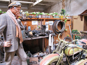 Full-scale model of a 1920s man in a workshop, contemplating a vintage Harley Davidson motorcycle.