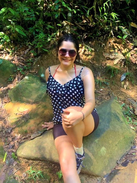 Trilha Praia do Sono em Paraty mulher jovem de cabelos pretos, oculos escuros, blusa preta com bolinhas brancas, short de corrida preto, tenis de caminhada marrom, sentada em uma pedra na natureza