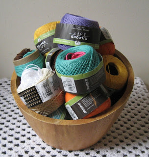 A wooden bowl full of 50 gram skeins of 4 ply cotton sits on a white crocheted granny stitch table cloth.