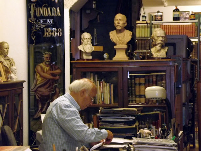 um senhor sentado numa mesa mexendo em vários papéis por entre estantes de livros e fotografias e um busto de um escritor