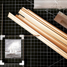Flatlay of a cutting mat, with lengths of miniature framing, a hobby saw, mitre box and gluing jig with a picture of a boat inside it.