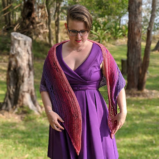 Photo of a white woman (Sarah) standing on a grassy space with trees, wearing a purple dress and a purple and pink crochet shawl.