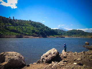 Peaceful Lake Water Dam Scenery With A Man Fishing Between Chunks Of Rock On A Sunny Day Titab Ularan North Bali Indonesia