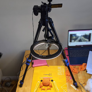 A camera set up on a tripod sitting on a dining table, the camera is pointed straight down to the table top which has a play mat with unassembled crochet orange frog plushie. Beside the setup is a laptop with Twitch stream blurred out.