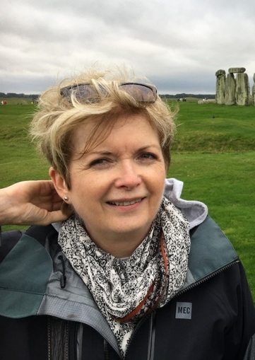 Doings in Bath: windblown hair with Stonehenge in the background woman in raincoat, holding down hair with Stonehenge in the background