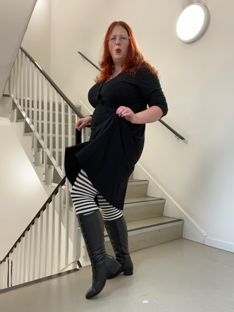 A red-headed woman in a black dress and black boots. She is raising her skirt to show off her black and white striped tights.