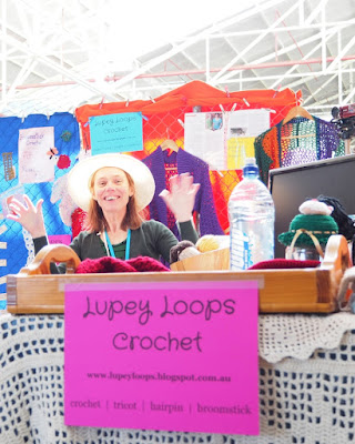 A low level view from the front of the display table (the short end of a rectangular trestle table). A pink printed A4 landscape sign says "Lupey Loops Crochet. www.lupeyloops.blogspot.com.au. crochet tricot hairpin broomstick". Behind the sign is an off-white filet crochet blanket.  Above it Jodie is waving both hands, being silly, and smiling. She is wearing a wide brimmed white hat and green long-sleeved top with the blue lanyard of the exhibitors pass around her neck.