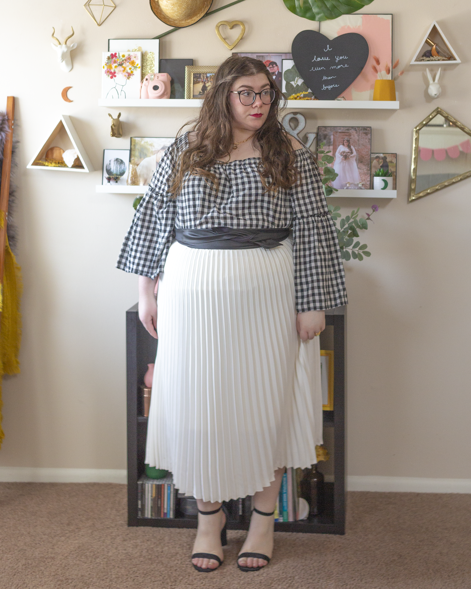 An outfit consisting of a black and white gingham off the shoulder with angel sleeves, tucked into a white pleated midi skirt and black strap heeled sandals.