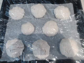 Baking tray with eight bread rolls on it. It is covered with cling film and is just starting to rise.
