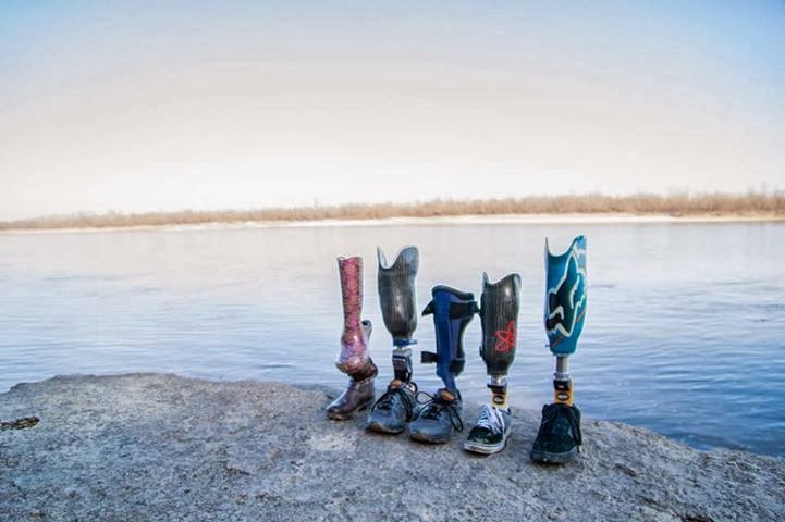 Five prosthetic legs, different sizes, different colors and patters, displayed in a row, on a flat rock surface next to a calm lake