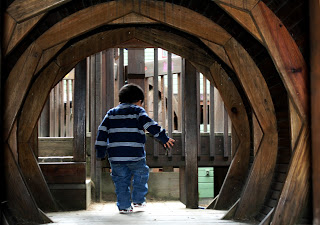 Boy walking away from the view through a play gym tunnel, exiting the tunnel into a larger play area
