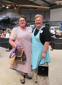 Two women dressed in 1940s outfits, standing in front of displays at a miniatures convention.
