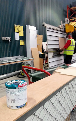 Cutting rea of a hardware store, with pieces of MDF and a test pot of paint in the foreground and an assistant cutting MDF in the background.