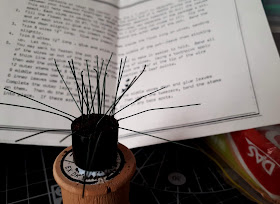 1/12 scale miniature plant kit with bare stems in a plant pot filled with 'soil' on top of a cotton reel, with a packet of air-drying clay to one side and a set of instructions behind.