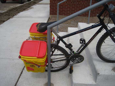 Bike chained to railing with two Tidy Cat kitty litter buckets used as panniers around the back tire