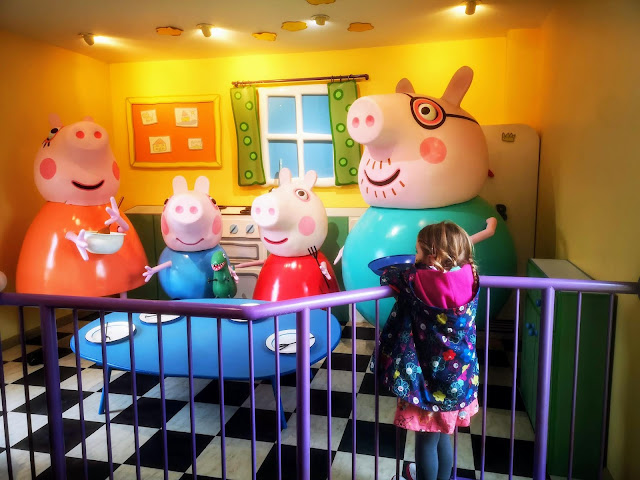 Image of a young girl stood at a metal fence looking out onto a well known scene from the Peppa Pig Cartoon. The scene features mummy pig, daddy pig, peppa pig and george pig as they make pancakes. Daddy pigs pancakes is stuck to the ceiling.