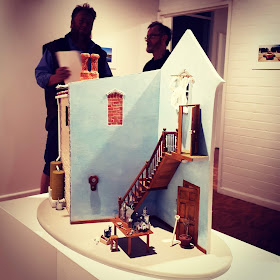 One-twelfth scale model of a two-story building on display in a gallery with teo people in discussion behind it.