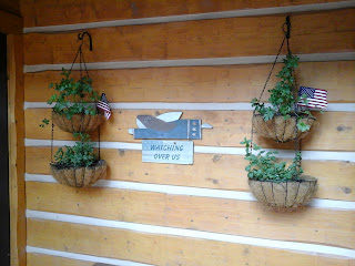 Exterior photo of 2 hanging baskets filled with herbs and small American flags.  Hanging on log siding and a red white and blue angel mounting on the siding betweend the hanging plants.