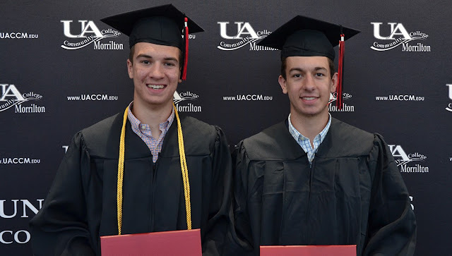 Two men wearing graduation robes and hats hold up diploma holders