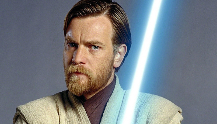 Imagem: o personagem Obi-Wan Kenobi, interpretado por Ewan McGregor, um homem branco com cabelos e barba curta e ruivas, em trajes Jedi brancos com detalhes marrrons e um sabe de luz azul e por trás um fundo cinza-claro.