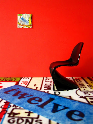 One twelfth scale modern miniature room with a giant map floor with the word 'twelve' across it, a modern black chair and a red wall with a small version of the floor print hung on it