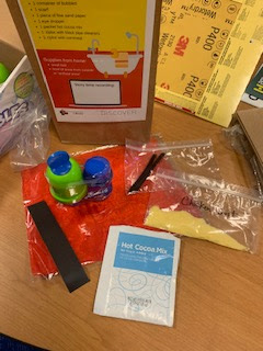 Closeup of various story time supplies in front of small paper bag with label, including bubbles, a scarf, hot cocoa mix, and bags of cornmeal and pipe cleaners
