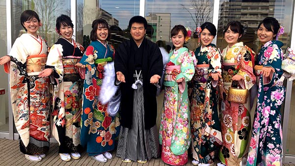 seven Japanese girls in full kimono are lined up in front of the camera, with a young man in hakata (traditional wear) in the center
