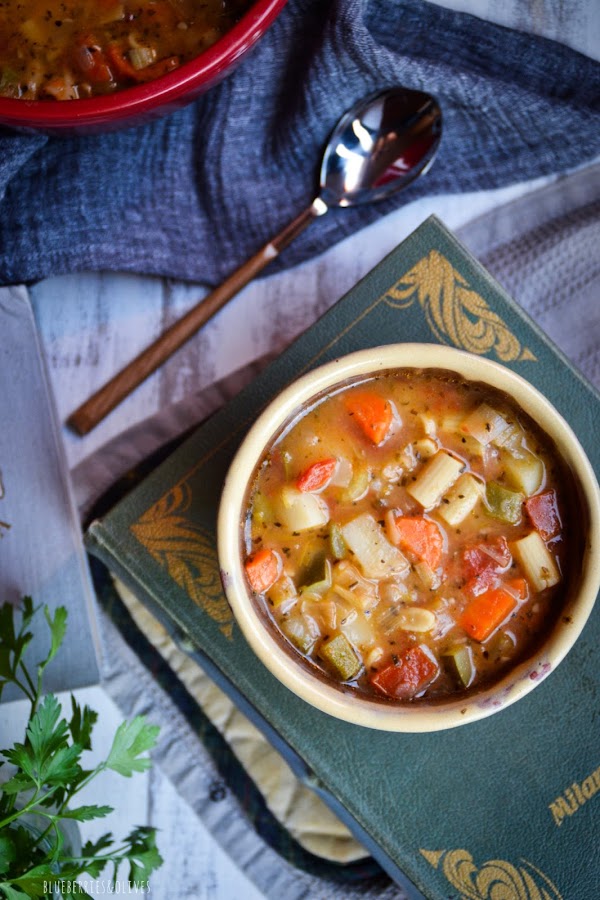 Olla cerámica roja llena con hoja laurel, bol cerámica con sopa, paño lino gris, libro cocina del mundo, cuchara mango madera,hojas perejil fresco