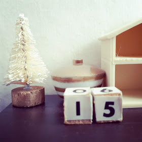 1/12 scale modern miniature tabletop scene in cream and brown containing a tiny bottlebrush Christmas tree, a lidded bowl with gold and white stripes, wooden blocks with the numbers '1' and '5' on them, and an empty dollshouse.