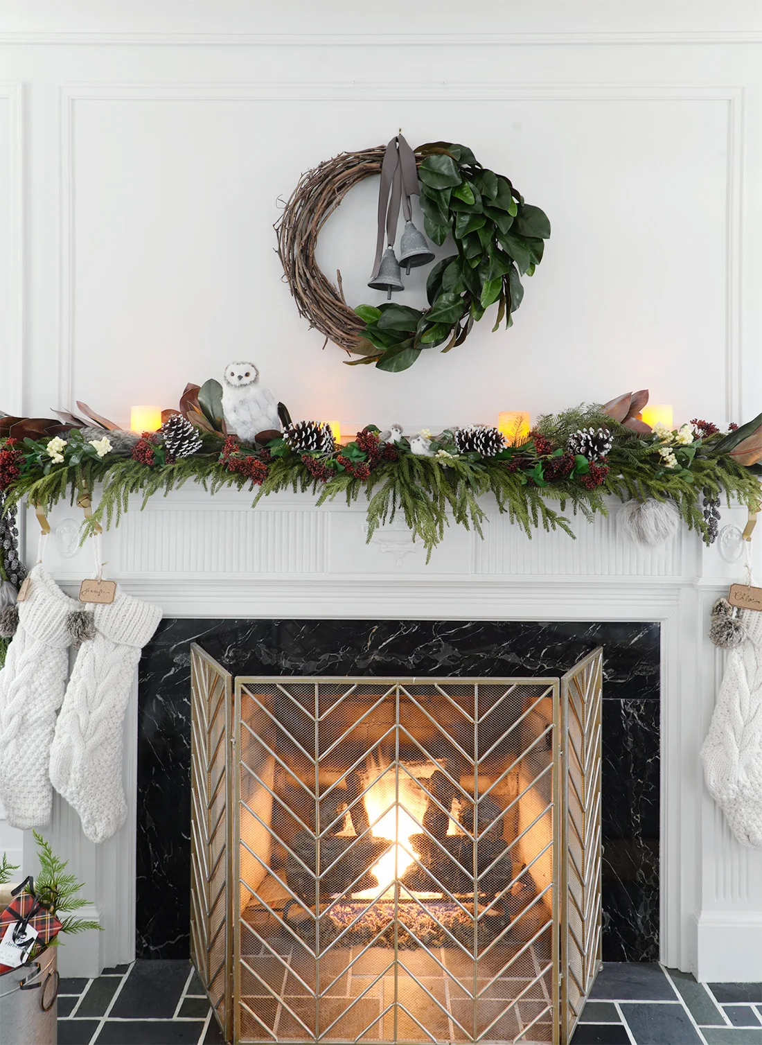 a faux christmas fireplace mantel garland with red seeded eucalyptus, a cedar garland and magnolia leaves. A vine and magnolia asymmetrical wreath with satin ribbon and metal bells. A brass chevron fireplace screen.