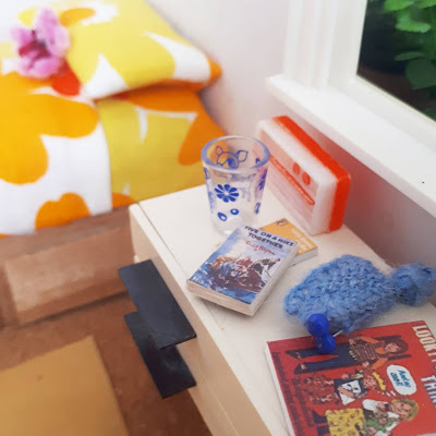One twelfth scale modern miniature 1970s child's bed and bedside table. The bed is dressed with bright flowery linen and the bedside table holds a flowery glass, an orange radio and a famous five book.