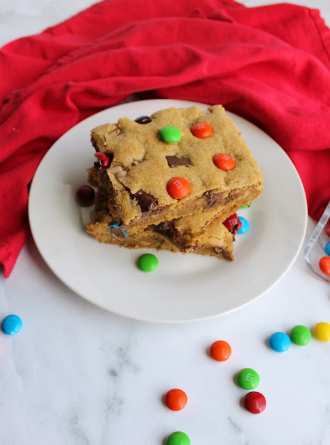 Chewy brown sugary blondies loaded with chocolate chunks, caramel undertones and topped with M&Ms. This big batch of bars is perfect for a party, carry-in or just because. looking down on a couple of pieces of chewy salted caramel m&m blondies on plate with candies scattered around