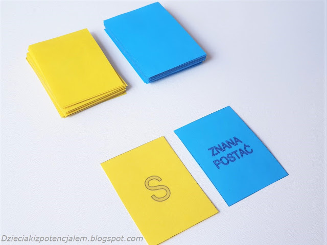 Na obrazku mamy karty w dwóch kolorach, żółtym i niebieskim, część kart leży w dwóch zakrytych stosach a dwie z kart leżą odwrócone, na żółtej jest litera s a na niebieskiej kategoria znana postać