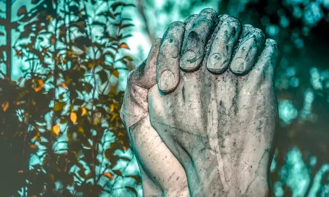 A imagem mostra duas mãos completamente sujas de barro cruzadas em gesto de oração.
