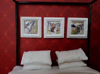 Three framed inchies of women from the 1950s with butterfly wings against a background of $10 Monopoly money on the wall above a one-twelfth scale modern miniature four poster bed with white sheets, blanket and pillows.