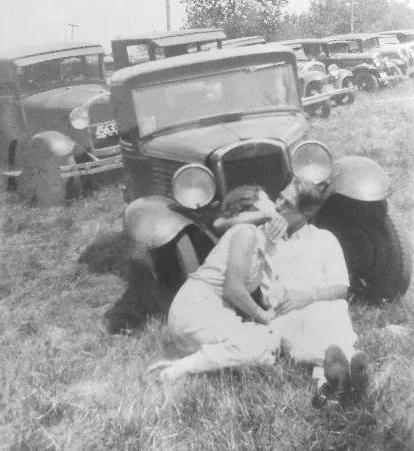 couple sitting in grass kissing in front of antique cars