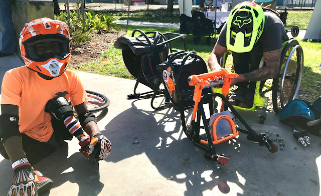 A man in a bright green, manual wheelchair with a matching helmet, is shown repairing another person's wheelchair. Its wheels are removed, and he is tinkering with the bear frame of the wheelchair. The wheelchair's owner is wearing a bright orange shirt and matching helmet, sitting on the ground and looking at the camera while his wheelchair is being repaired.