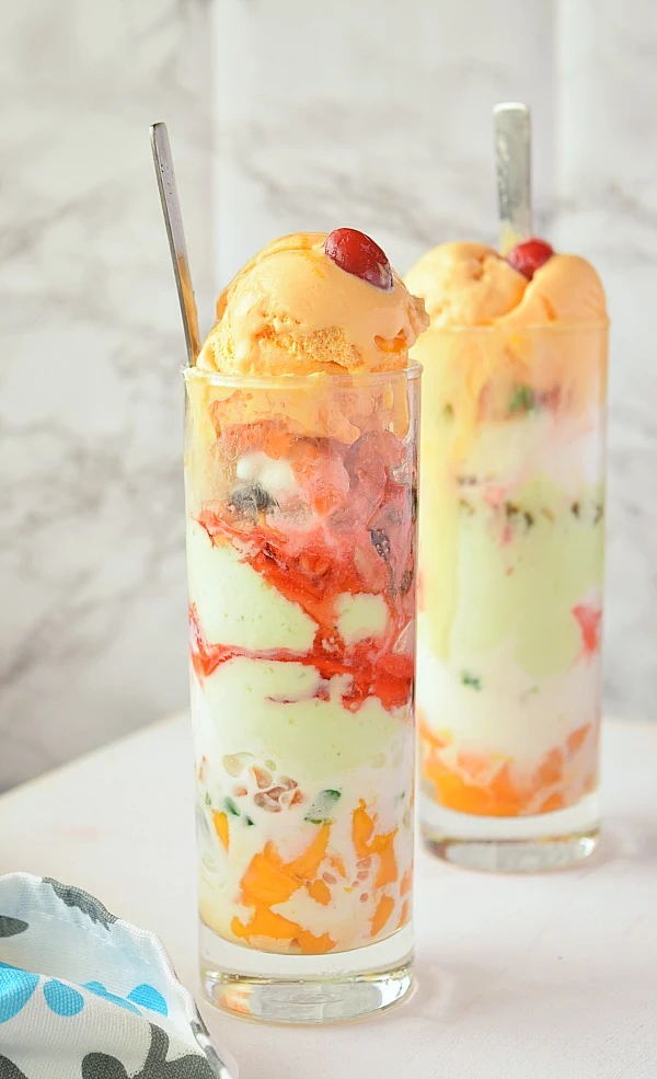 tall glass of Sundae Ice Cream Gadbad with layers of Mango,Pistachio and Vanilla icecream,mango chunks and fruits