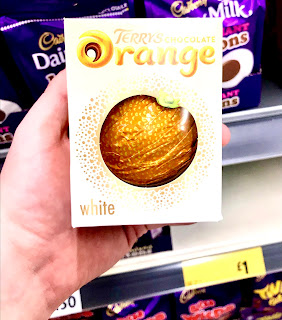 A square white box with Terrys in black cursive font and chocolate in black bold font and orange in orange bold font with a circle in the middle of the box where there is a spherical white chocolate ball that resembles an orange via having semicircular segments in a bright orange wrapper with white underneath this on the box in small white font on in front of a row of bright purple rectangular pouches on a light brown rectangular shelf on a bright background