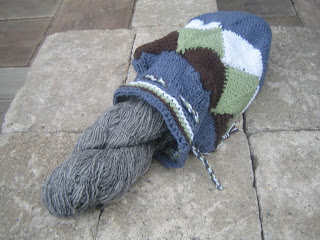This shows a drawstring bag made primarily of joined mitered squares. The top has eyelets with a twisted cord threaded through them, and a skein of yarn is partially out of the open bag.